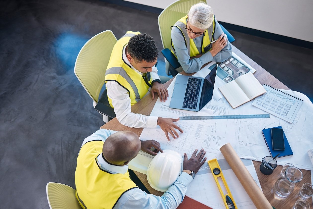 Top‑down view of a team meeting with a laptop and printed floor plans during construction planning.