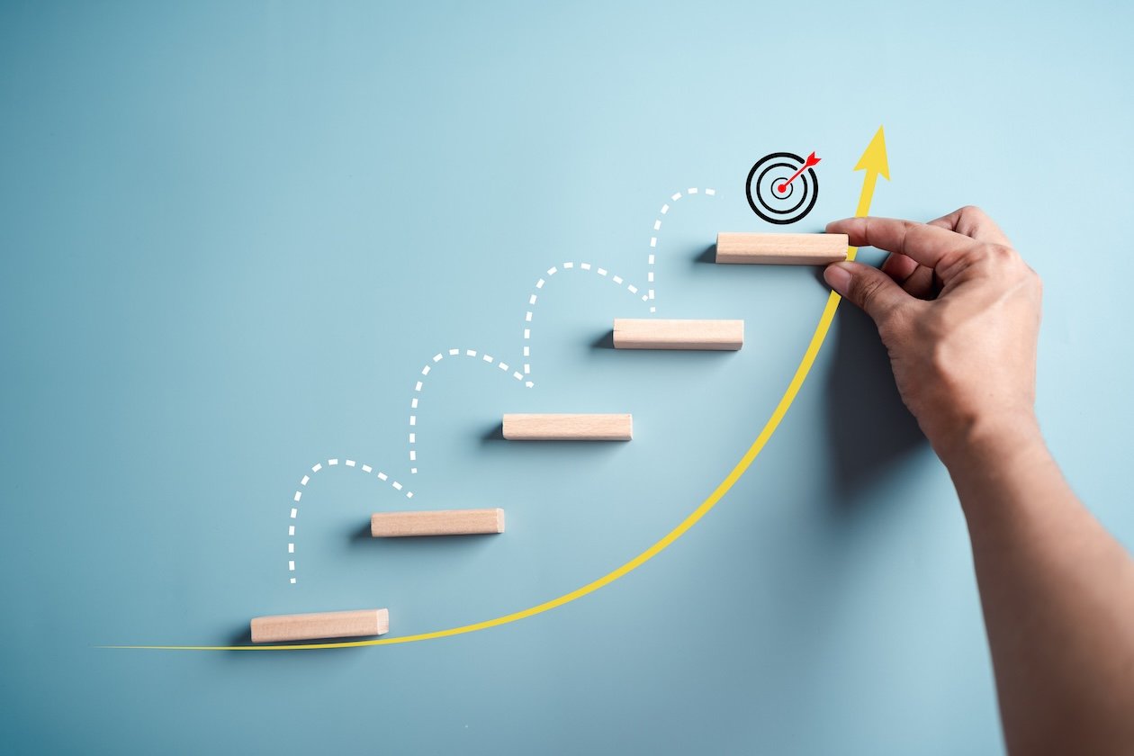 Hand placing wooden steps upward along a rising curve toward a target symbol, representing business growth, progress, and operational improvement