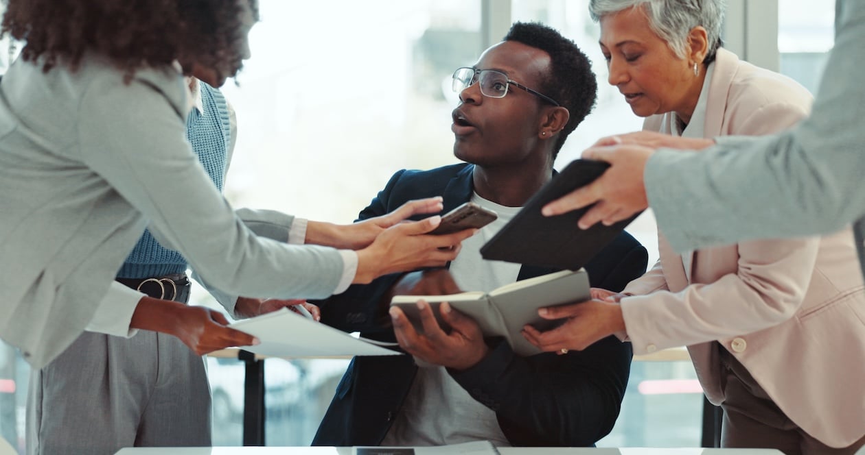 Overwhelmed office manager juggling phone calls, documents, and digital tasks while coworkers demand attention, illustrating scheduling chaos and communication overload in a busy workplace.