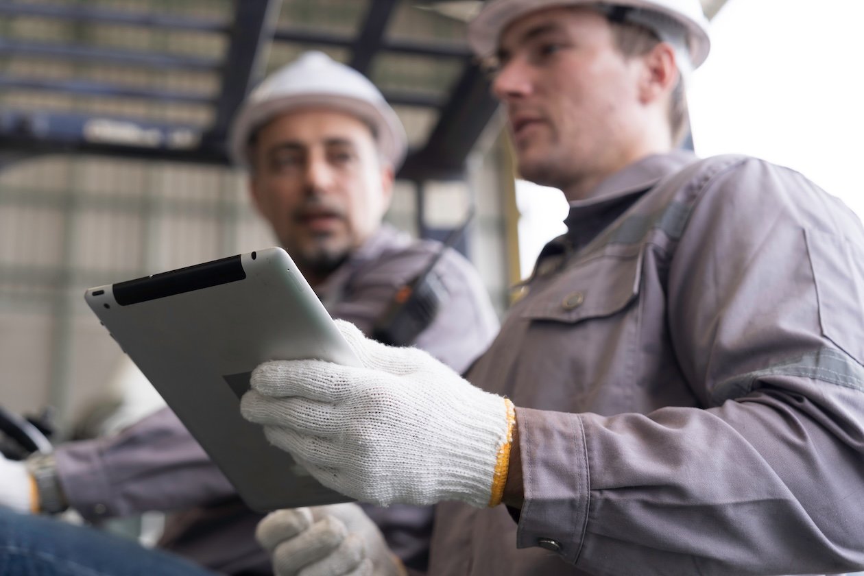 Engineers in safety gear reviewing project information on a tablet at an active worksite, illustrating connected job data, real-time collaboration, and coordinated restoration project management across teams and workflows.