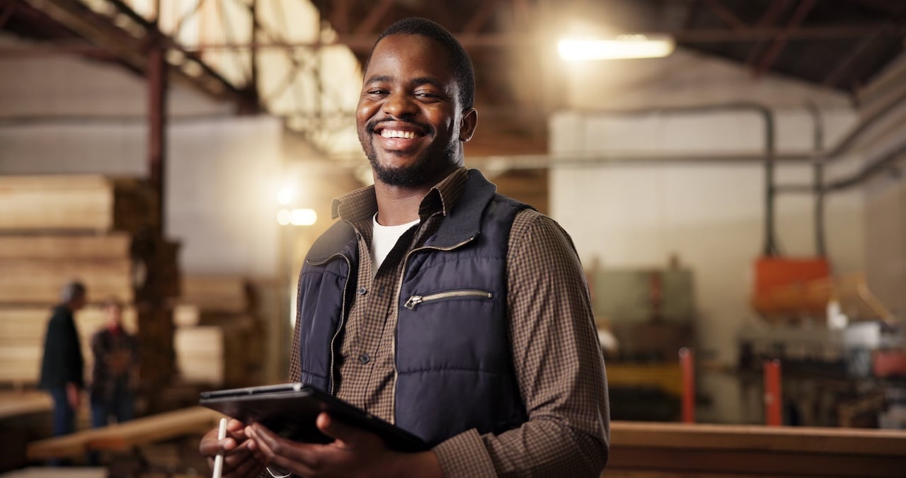 Smiling male carpenter holding a tablet in a woodworking shop, checking furniture designs and inventory for manufacturing projects.