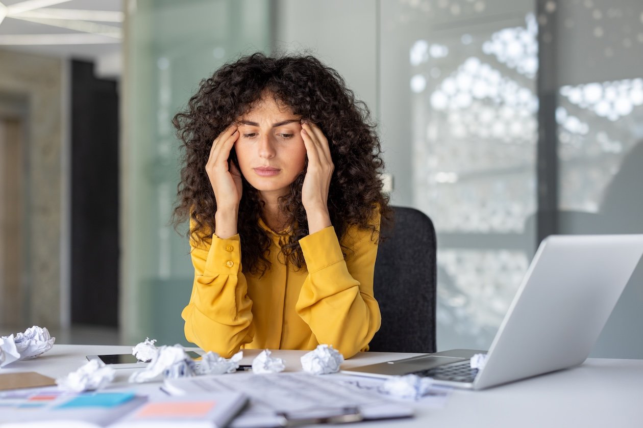 Stressed businesswoman sitting at a cluttered desk surrounded by scattered paperwork, reports, and a laptop, illustrating fragmented information, disconnected workflows, and operational visibility challenges that can lead to misleading profit margins in a growing business.