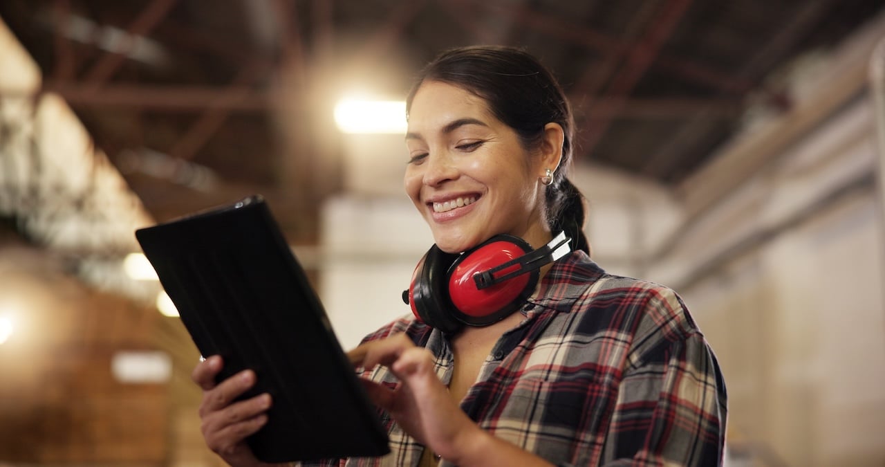 Female carpenter smiling while using a tablet in a woodworking shop, reviewing designs and inventory for furniture manufacturing.